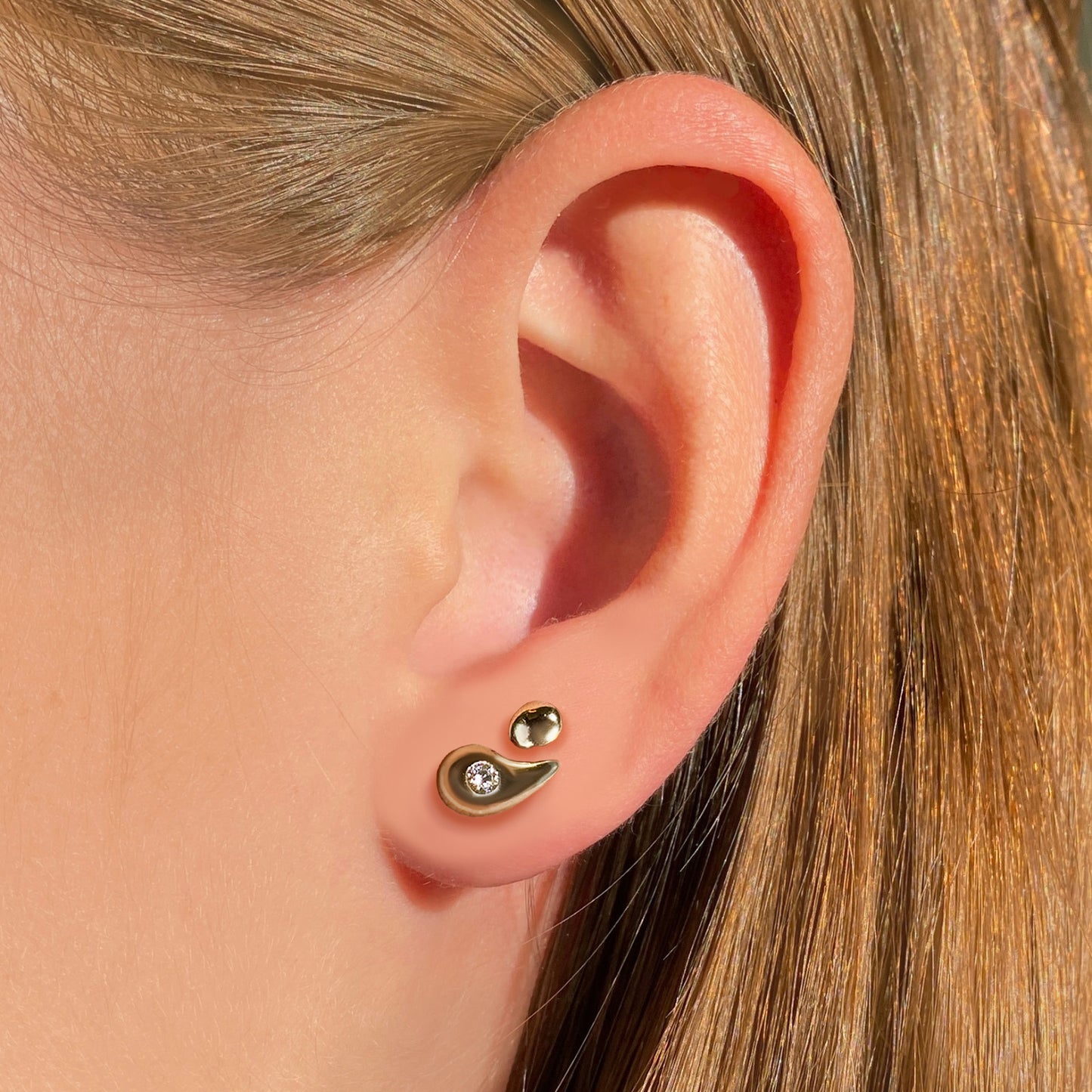 A Person wearing a teardrop-shaped yellow gold earring with a round gold earring