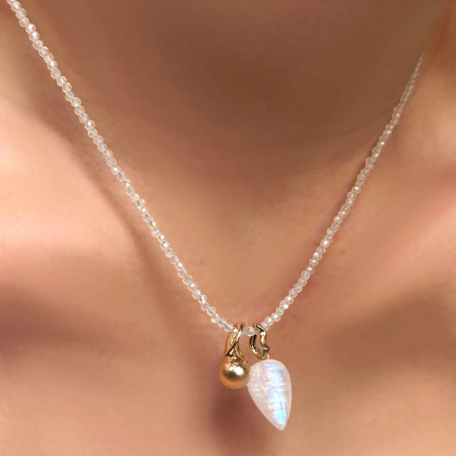 A moonstone drop charm with a 14K yellow gold ripple bale hangs from a moonstone slim gemstone necklace, along with a 14K recycled yellow gold mini molten drop charm.
