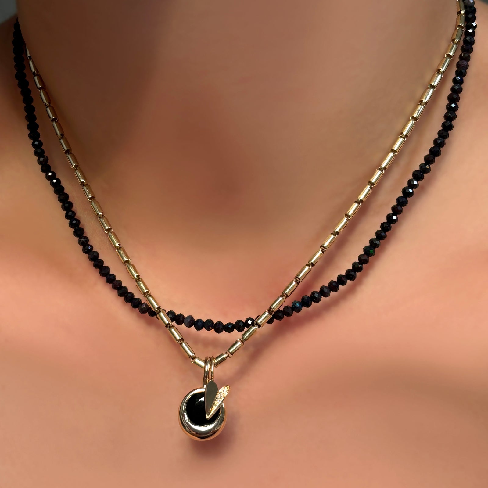A model wears a sparkling black faceted opal necklace, paired with a yellow gold sprinkle chain, a half pavé 14K yellow gold heart charm,  and a black onyx round molten solitaire charm with a 14K yellow gold bezel.