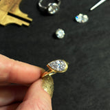 Yellow gold molten solitaire ring with tipped pear shaped diamond being held by a model with gold nail polish against a black background with tools and diamonds.