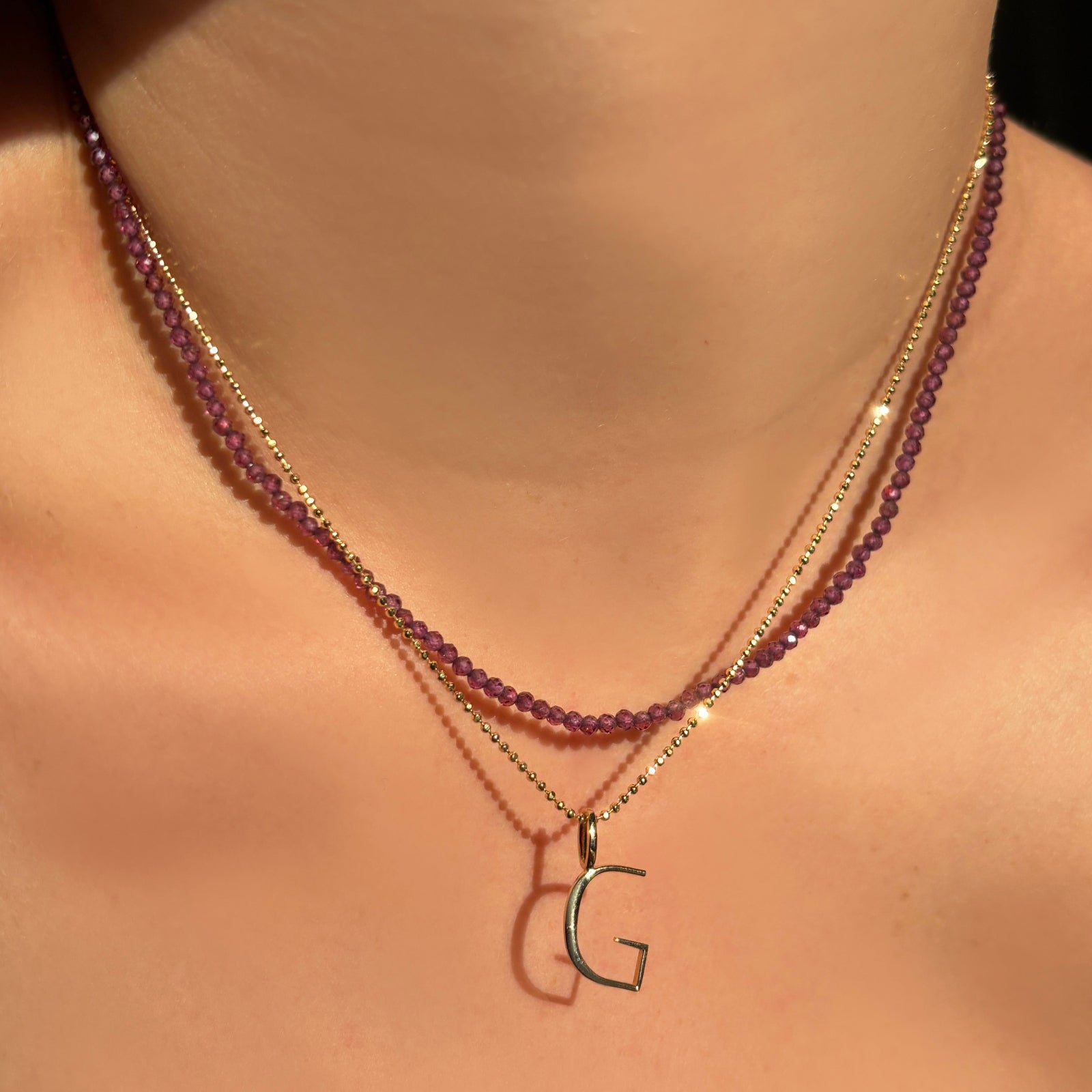 purple garnet slim gemstone layered with a diamond cut bead chain with letter "G" charm on a model's neck.