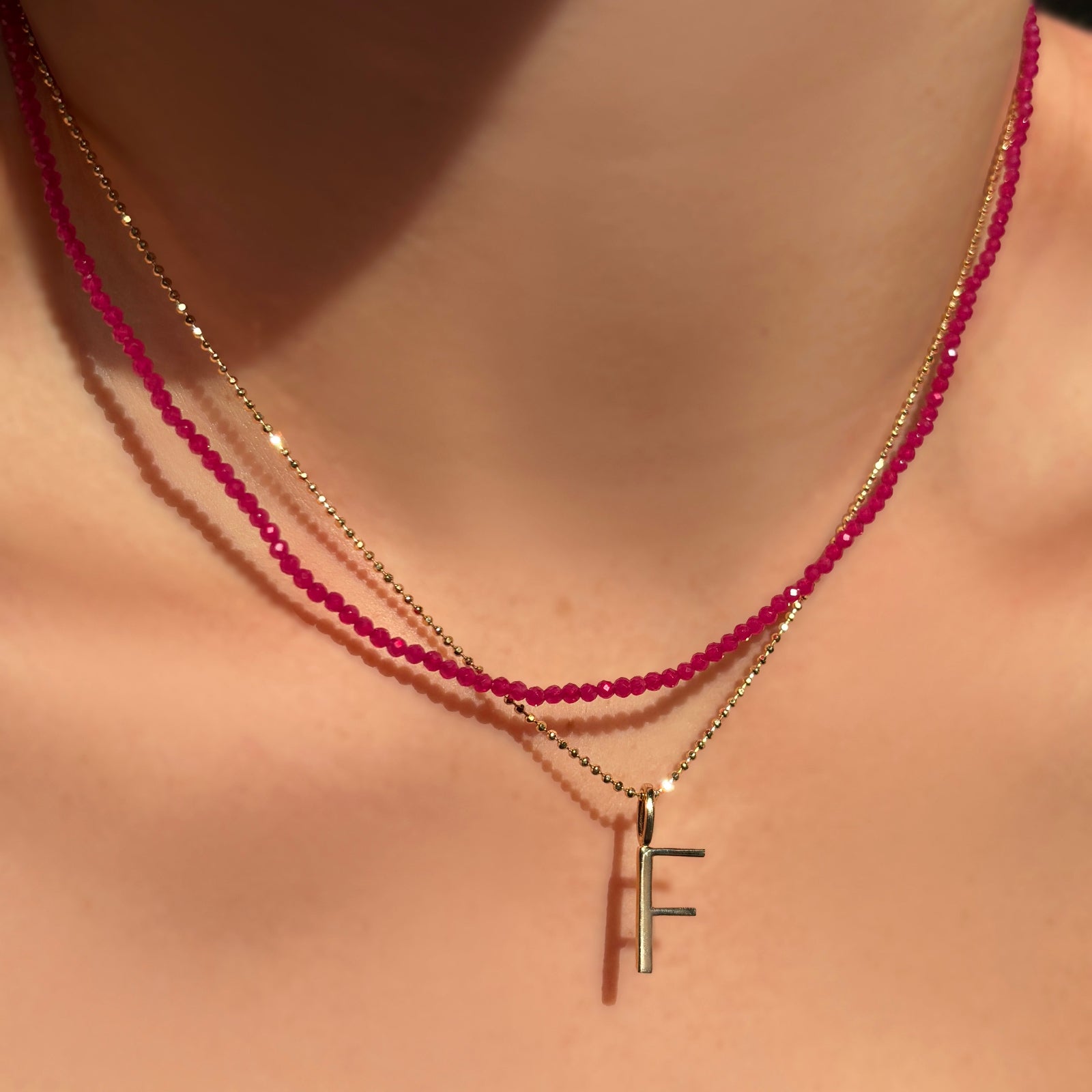 Faceted ruby slim gemstone necklace layered with a diamond cut bead chain with letter "F" charm on a model's neck.