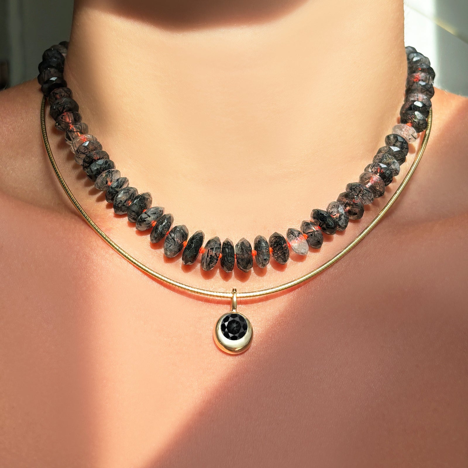 Necklace with rutilated quartz beads strung on a neon red thread layered with a gold chain and round yellow gold charm set with a black onyx stone.