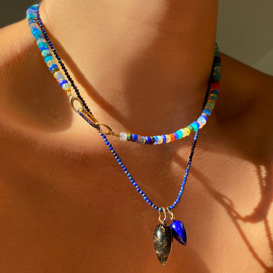 Shimmering beaded necklace made of faceted opals in shades of fiery blues, greens, teal, black, orange, and clear on a gold linking ovals clasp. Styled on a neck layered with a small sapphire charm lock and two acorn drop charms
