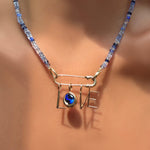 A denim faceted sapphire necklace with a recycled 14k gold molten safety pin in its evolution clasp, dropping letter charms and a round molten sapphire solitaire charm spelling out LOVE on a model's neck.