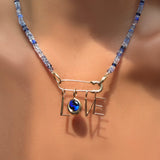 A denim faceted sapphire necklace with a recycled 14k gold molten safety pin in its evolution clasp, dropping letter charms and a round molten sapphire solitaire charm spelling out LOVE on a model's neck.
