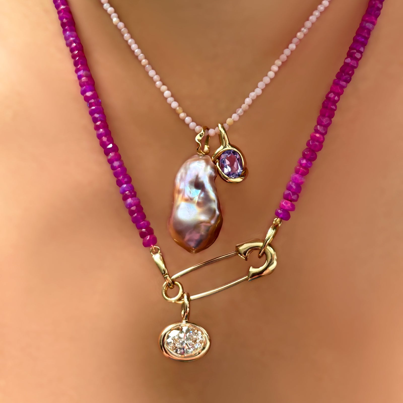 A fuchsia faceted opal necklace clasped with a 14K yellow gold safety pin charm lock from which hangs a 14K recycled yellow gold molten oval charm set with an ethical lab grown diamond. It is layered with a slim pink opal necklace from which hangs a baroque pearl charm with ripple bale and a 14K molten knot charm with amethyst. 