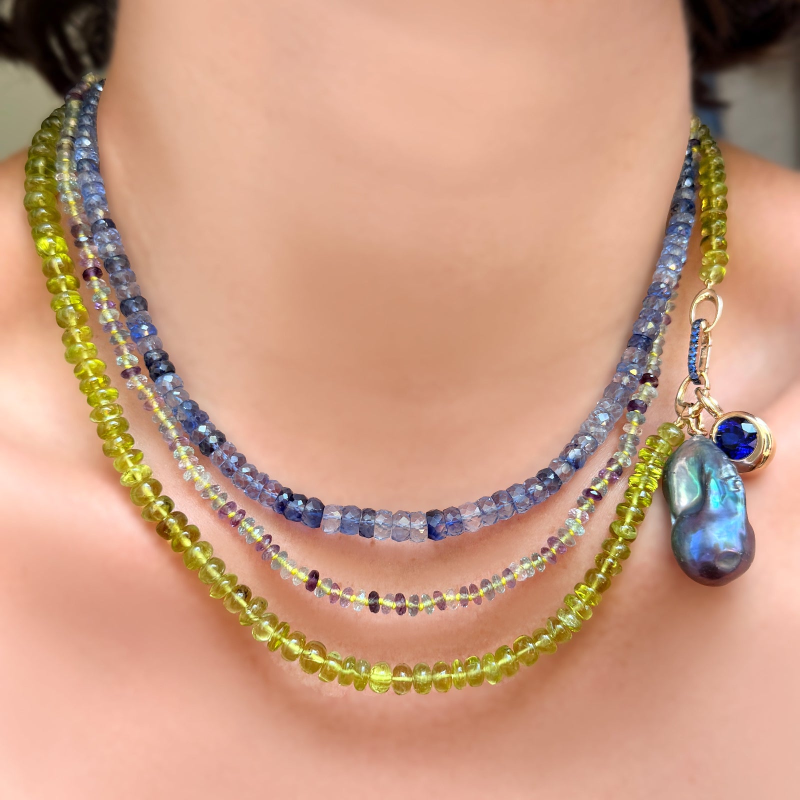 Faceted denim sapphires layered with faceted fluorite on neon thread over a strand of peridot rondelles with a storm baroque pearl charm, molten solitaire charm with ethical sapphire, and small sapphire charm lock on a model's neck.