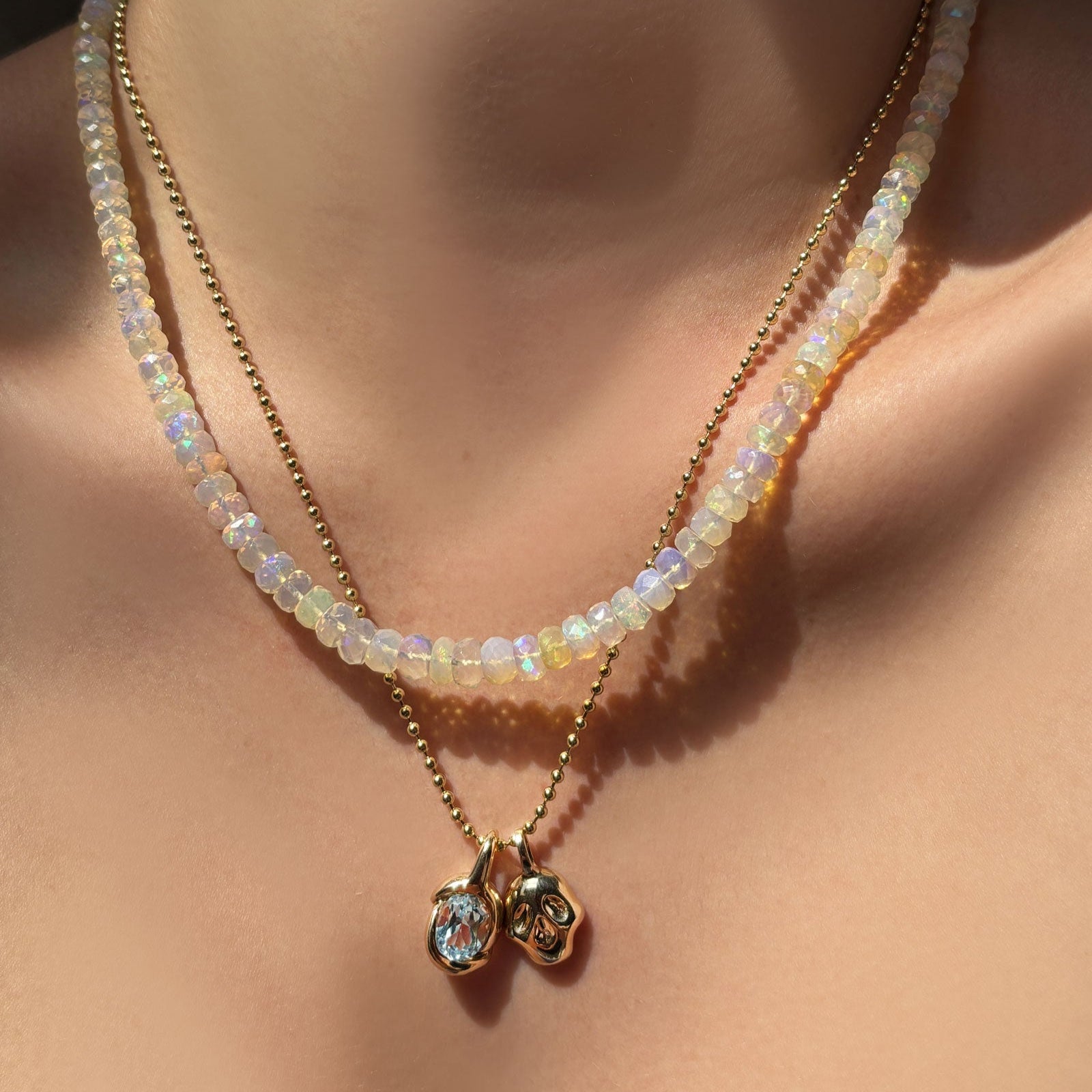 Supermoon faceted opal necklace layered with a gold bead chain, molten knot charm with blue topaz, and molten skull charm hanging on the neck.