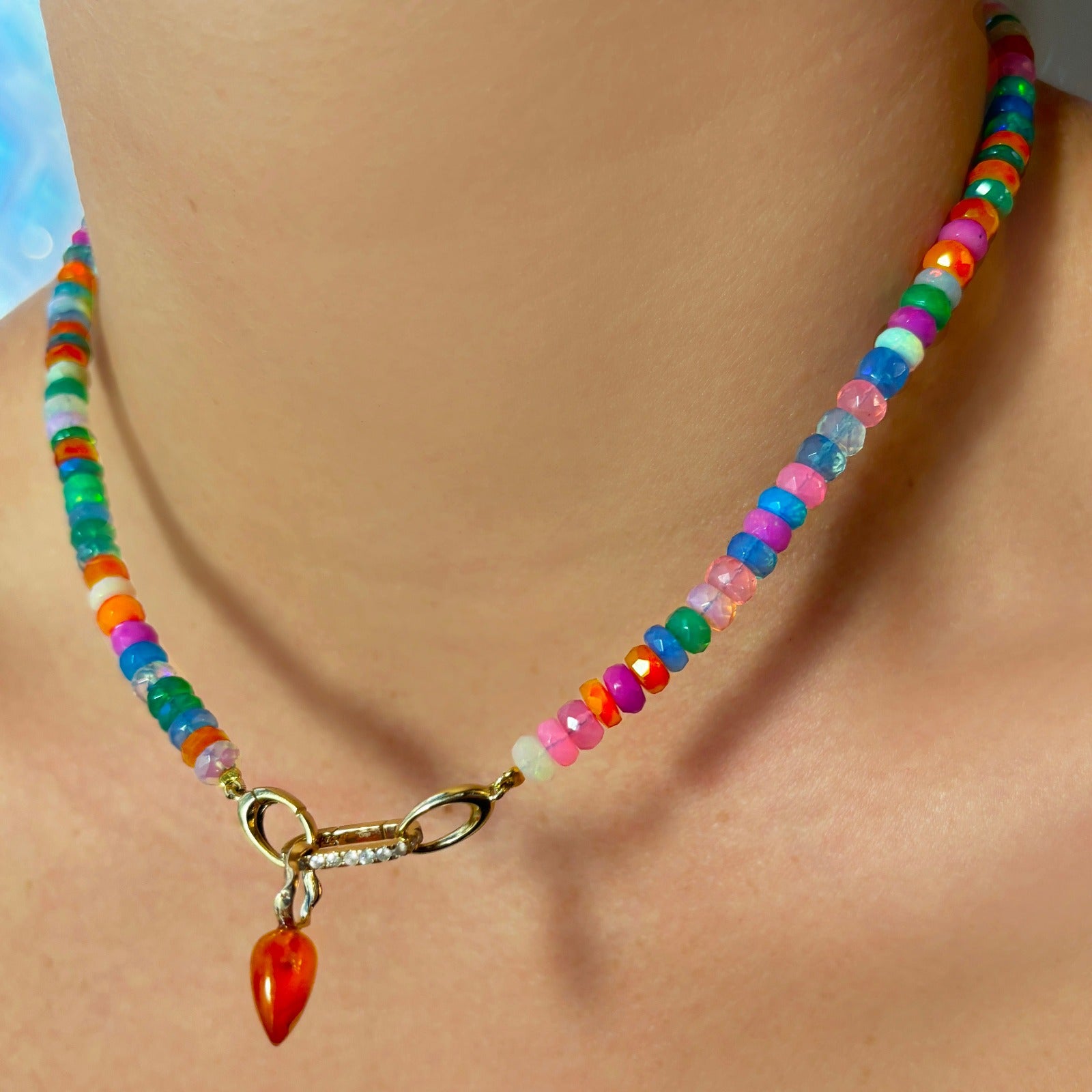 Shimmering beaded necklace made of faceted opals in shades of fiery blues, yellows, teal, and purple on a gold linking ovals clasp. Styled on a neck layered with a small diamond charm lock and acorn drop charm.