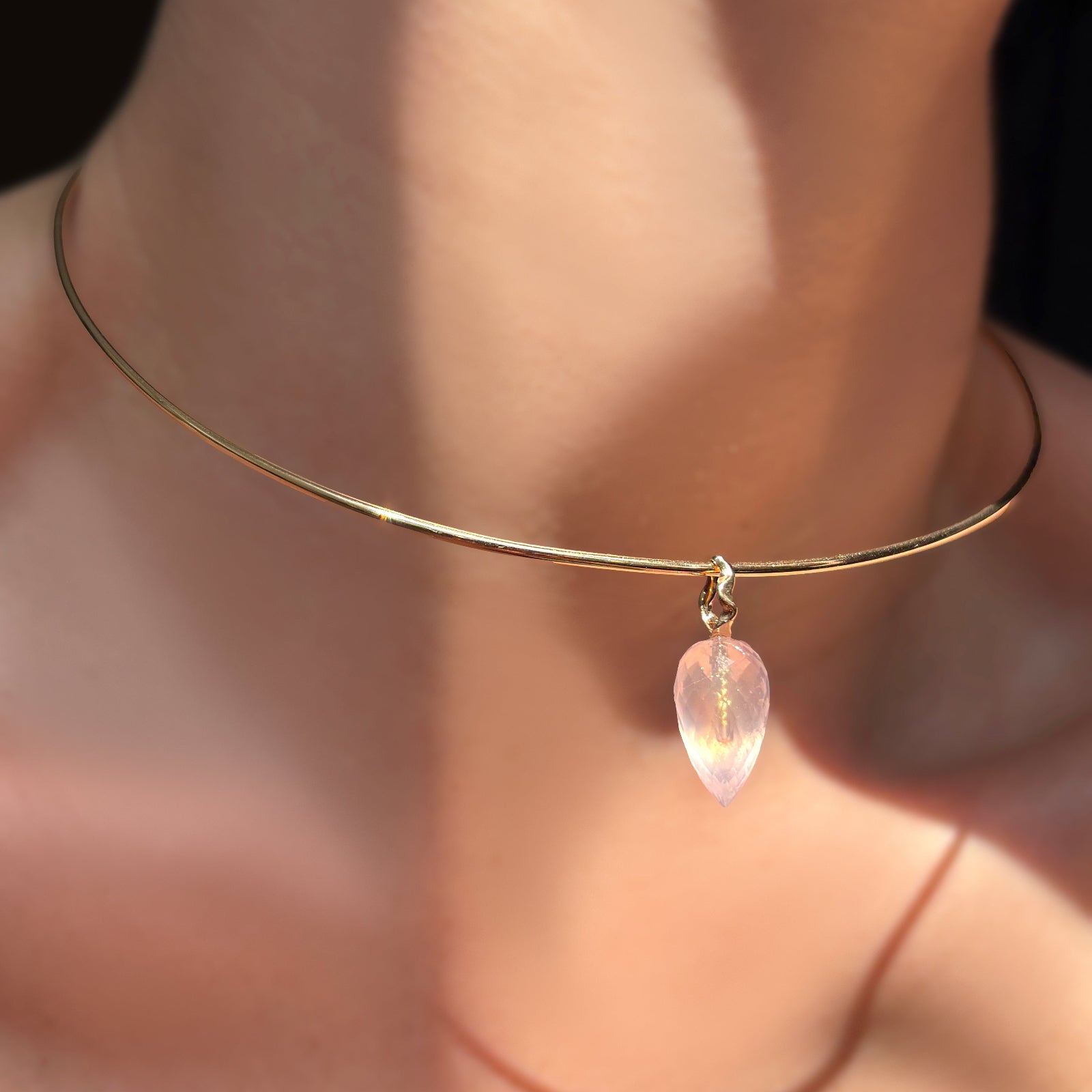 Faceted rose quartz drop charm on 14k gold ripple bale hanging from a wire choker on a model's neck.