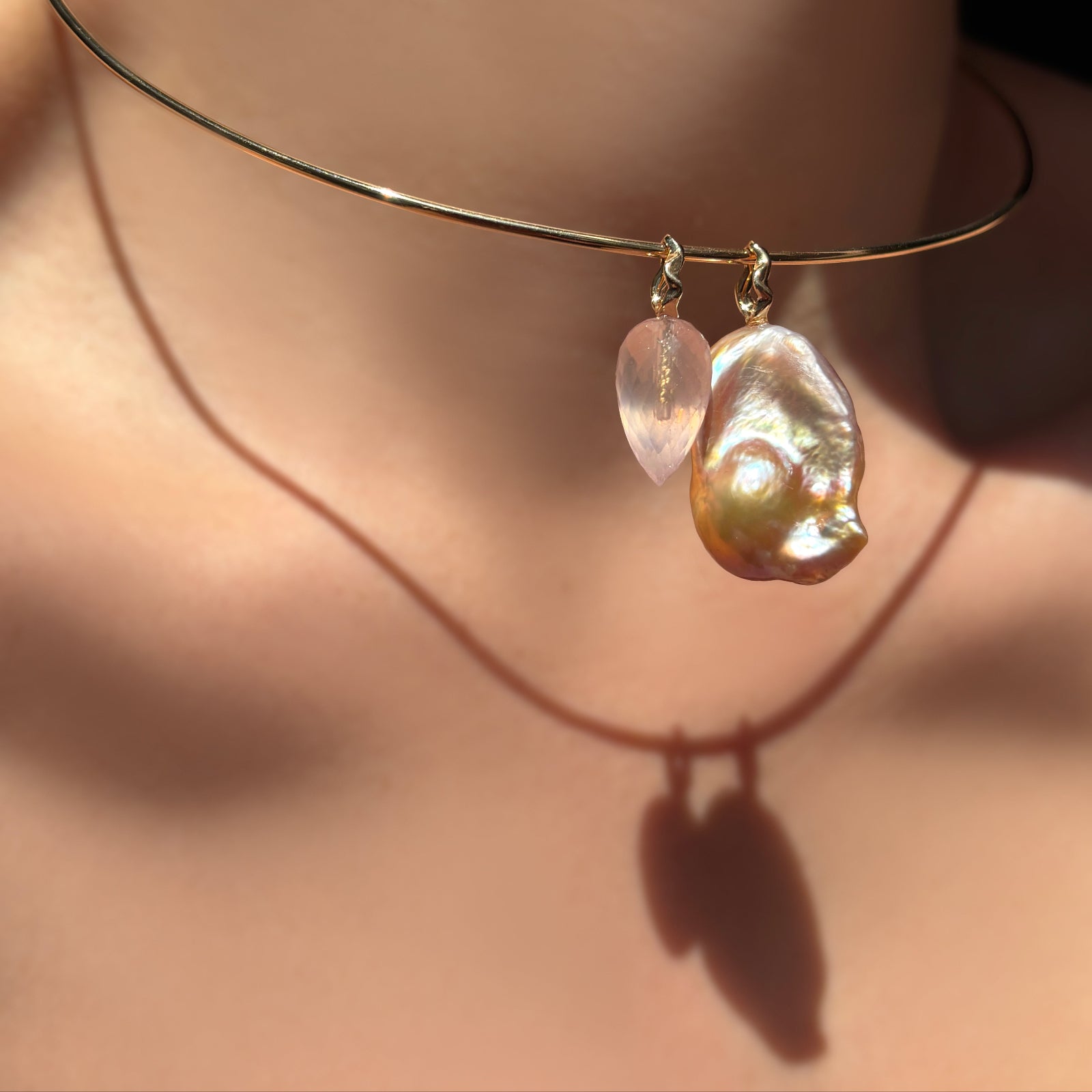 14k gold wire choker on a model's neck with a faceted rose quartz drop charm on ripple bale, and blush baroque pearl on ripple bale.